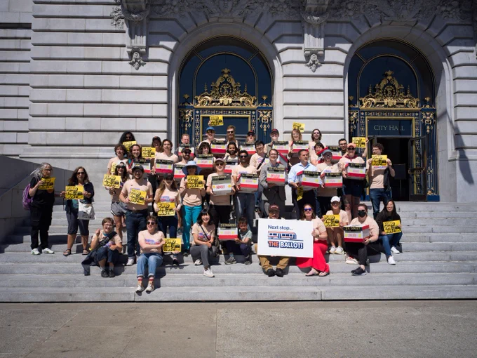 40 volunteers holding boxes painted like Muni buses and Fund the Bus signs in front of SF City Hall