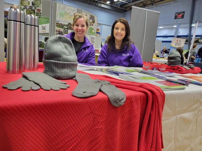 Jo and Katie sot behind a table with leaflets, hats, socks and gloves and flasks
