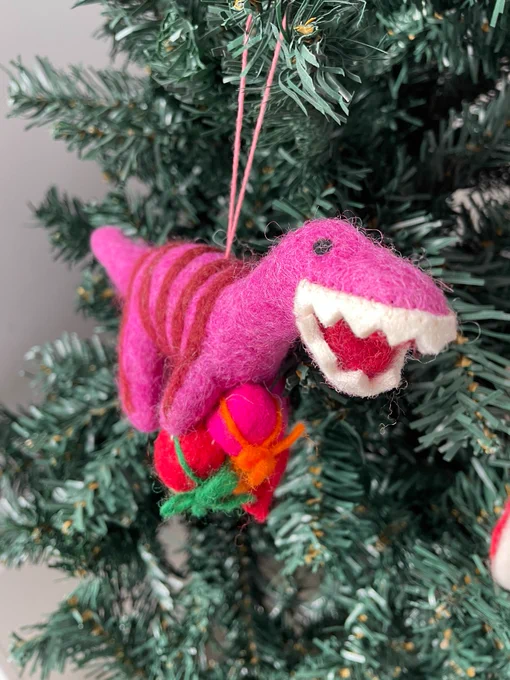 A pink felt dinosaur smiling with white teeth, holding two presents, hanging on a green Christmas tree