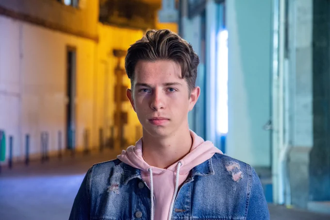 Young man looking directly into the camera with a neutral expression. Wearing a denim jacket and a pink hoodie.