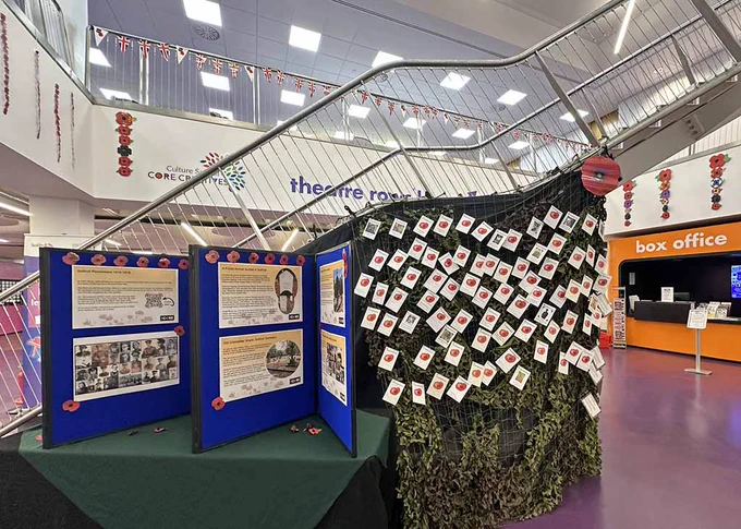 A photograph of the foyer at The Core showing a staircase with a table-top display in front and cargo netting covering the under stair area. Affixed to the netting are small cards bearing a picture of a poppy and details of individuals commemorated