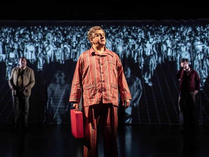 Person standing on stage in striped pyjamas holding a red ministerial box, with a black and white photo of a crowd projected in the background.