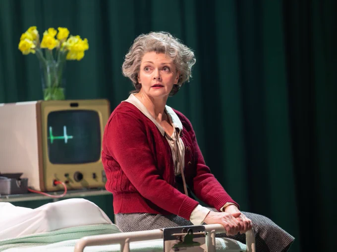 A person is seated beside a hospital bed, looking worried. They have short, curly grey hair and wear a red cardigan over a blouse paired with a checked skirt. The stage setting includes a green backdrop and a bouquet of daffodils. 