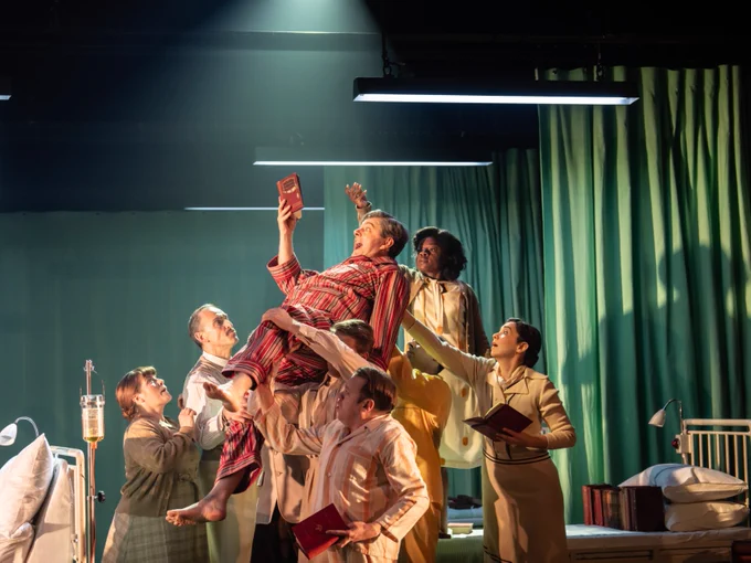 A dynamic theatrical scene with multiple performers lifting up a central figure holding a book, set against a stage backdrop with dramatic lighting.
