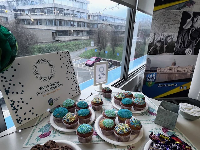 Table of freshly baked fairy cakes, topped with blue and green decorations, which complement the World Digital Preservation Day poster board, and the UCD Digital Library banner. View of the Newman Building out the window.