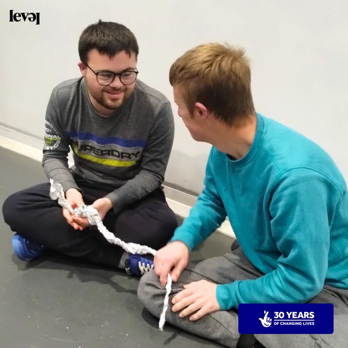 Two people sit on the floor in a room. They are making what looks like a snake out of tinfoil. They look mischievous and are having fun!