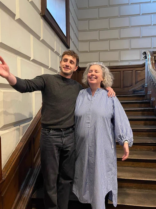 Linda smiling for a photo with Paul Mescal. They are standing on brown wooden stairs.