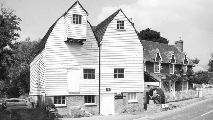 Haxted Watermill, a double fronted white weatherboard building with a small waterwheel to the right.