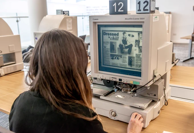 A person sits at a table using a microphiche machine. On the screen appears to be a digitised double page magazine spread with the headline: 'Dressed to chill'.