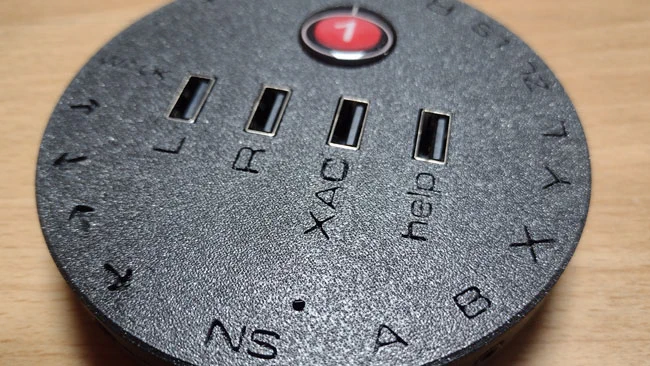 Black circular interface with OneSwitch sticker. 4x USB sockets on the top labelled L, R, XAC, help. Around the edge, A,B,X,Y, L and so on.