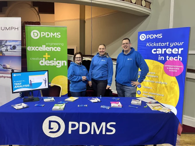 Three people in PDMS jumpers posing for a photo behind a PDMS stall at a careers event