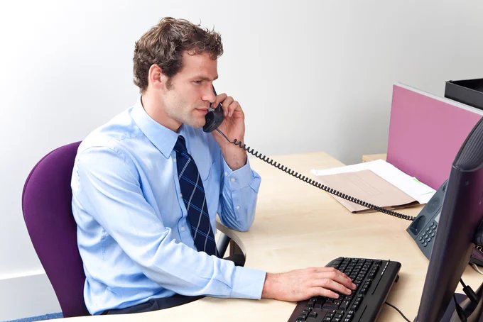 Stock image of clerical officer at work, viewing a PC monitor with a telephone in one hand and the other on PC keyboard. 