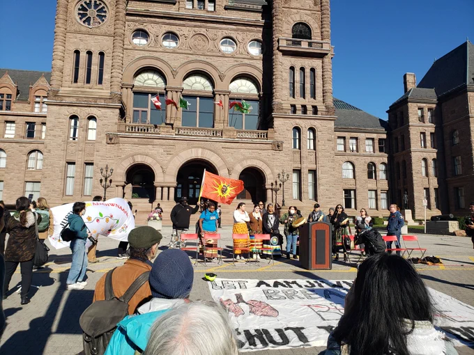 Outside the Queens Park building in Toronto on a sunny day, a few Indigenous people are drumming in a line. There is a wooden lectern and a few banners about Aamjiwnaang First Nation, and a red Indigenous warrior flag.