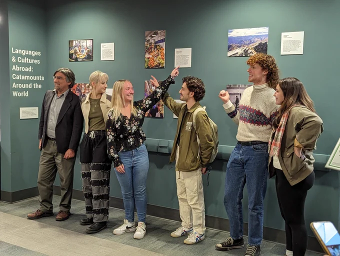 Five students stand in front of a photo exhibit smiling and pointing at their photos. Their professor stands next to them smiling. 