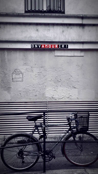 Mur d'un bâtiment. Des lettres sont collées et forment la phrase ONVAMOURIR ! tout attaché. Les lettres sont blanches sur fond noir sauf les lettres AMOUR qui sont sur fond rouge. Plus bas, un graffiti représente une plaque de rue avec le nom Rue Bite Chatte. Au premier plan devant une partie striée du mur : un vélo avec un panier à l'avant est attaché à une balustrade. 
