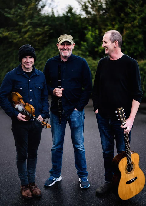 Michael McGoldrick, John McCusker and John Doyle stood outside holding their instruments, with trees and bushes in the background behind them. 