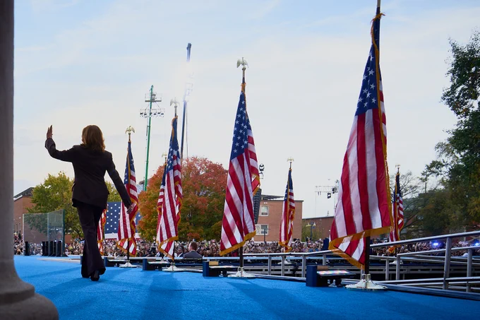 Vice President Kamala Harris waving to a crowd while walking on stage to deliver her concession speech. There are American flags displayed down the stage and there is a large crowd in the background.  