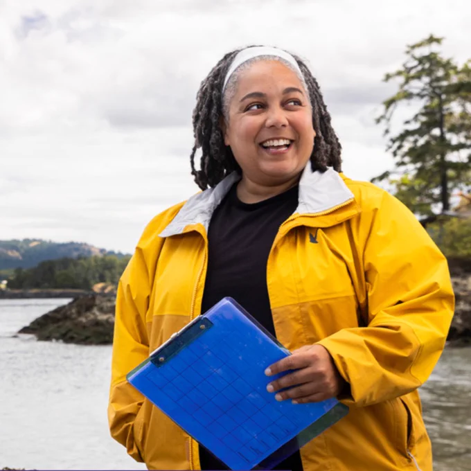 Photo of Chris Mantegna in a yellow coat holding a blue plastic clipboard next to Puget Sound with trees and islands in the background.