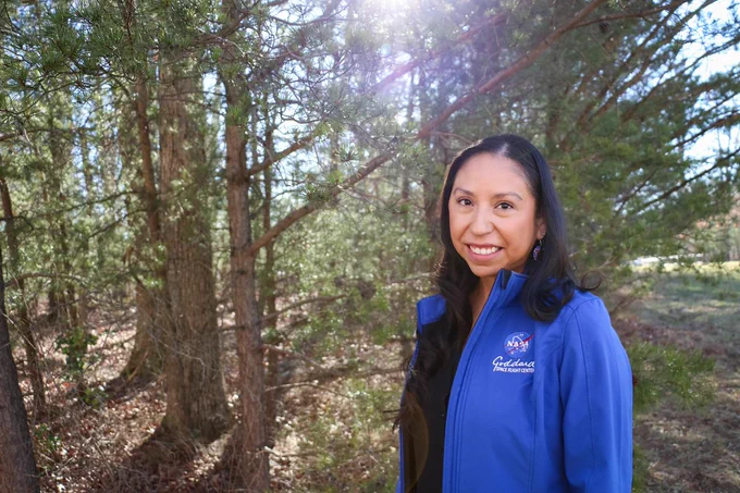 Miranda Meyer, a member of the Eastern Band of Cherokee Indians from the Birdtown Community of North Carolina, poses for a portrait amid the trees at NASA’s Goddard Space Flight Center. She has long, dark hair and is wearing a bright blue jacket with the NASA and Goddard logos on it. Credit: NASA/Thalia Patrinos