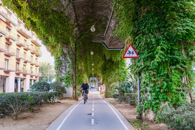 Pedestrians, cyclists, scooters, and joggers travel along a pathway underneath a concrete railway viaduct in Girona featuring climbing plants, street art, and overhead lighting.