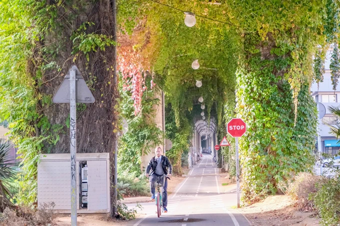 Pedestrians, cyclists, scooters, and joggers travel along a pathway underneath a concrete railway viaduct in Girona featuring climbing plants, street art, and overhead lighting.