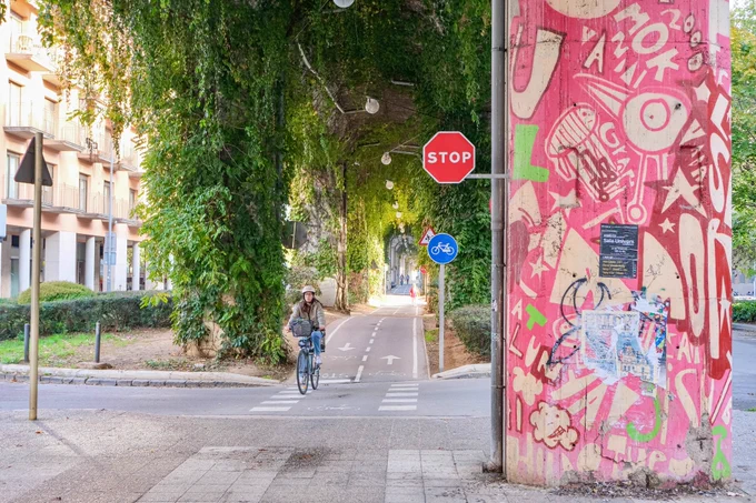 Pedestrians, cyclists, scooters, and joggers travel along a pathway underneath a concrete railway viaduct in Girona featuring climbing plants, street art, and overhead lighting.