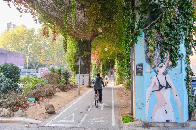 Pedestrians, cyclists, scooters, and joggers travel along a pathway underneath a concrete railway viaduct in Girona featuring climbing plants, street art, and overhead lighting.