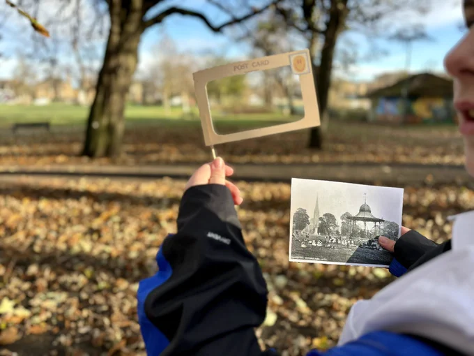 Photo taken in an autumnal park with bare trees and yellow and brown leaves on the ground. A  young person’s hands can be seen in the foreground, the right hand holding a black and white historic photograph of the park, the left hand holding up an A6 sized frame.