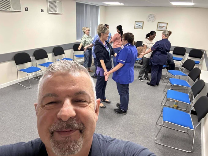 Trainer Pat taking a selfie, NHS staff in the background learning physical skills. conflict resolution training. 