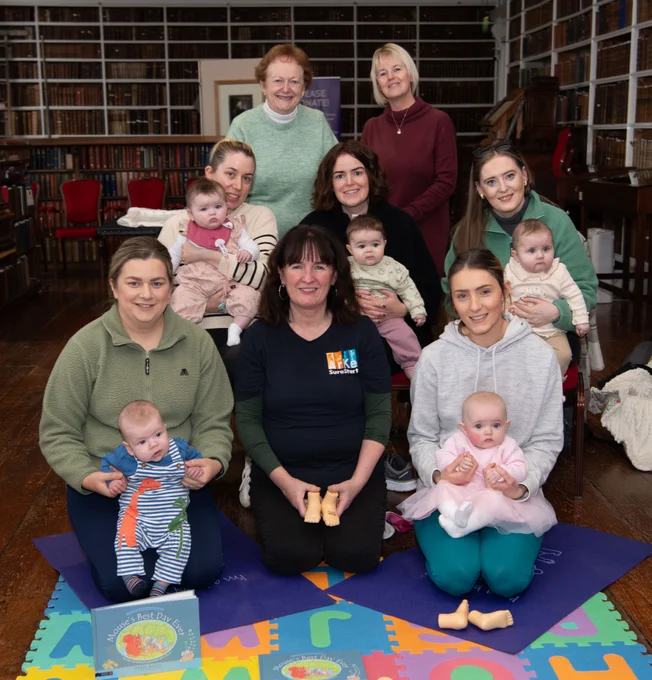 Eight women and five babies seated and standing in an historic library on a playmat.