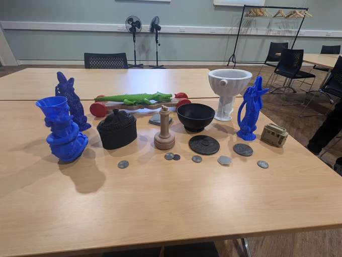 [17 3D printed objects in varying sizes. They are different colours including blue, white, and black. There are intricate animal design jugs, a large replica of a Burmantofts toilet and a handful of coins and medals.]   