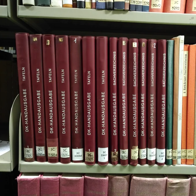 foto einiger exemplare der "DK-handausgabe" mit tafel- und sachverzeichnis-band, rot eingebunden, in einem bibliotheksregal
