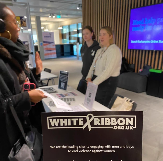 Image shows a stall with female students talking. In the foreground is a card that reads WhiteRibbon.org.uk 