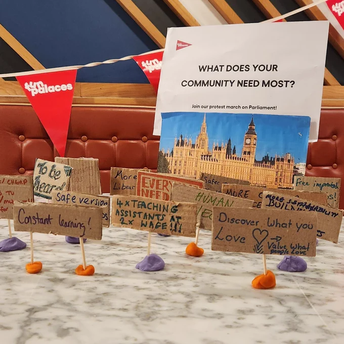 A miniature cardboard placard protest in front of a photo of the Houses of Parliament.