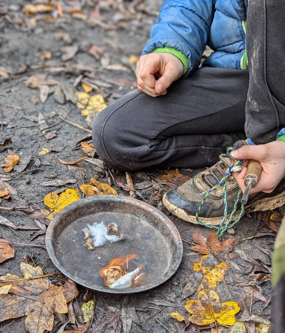 Child's legs, feet and arm, kneeling on a leafy floor by a metal tray with a small fire in it. The child holds a fire steel with a blue and yellow string.