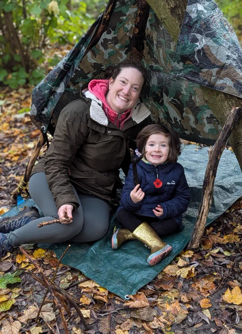Adult and child in a den. Adult wears dark green coat over a pink hoody and child is wearing navy coat and gold sparkling wellies. Den is made from sticks and a tarpaulin and they sit on a tarpaulin mat