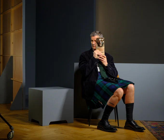 Des Hamilton poses with his BAFTA Scotland Award for Outstanding Contribution to Craft. He is sitting down in a chair and holding the trophy up to cover one side of his face. He is wearing a green and blue tartan kilt and suit jacket. 