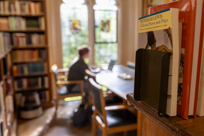 Interior of a college library with a person seated at a table reading near a window with stained glass. A book titled 'Wyndham Lewis: Collected Poems and Plays' is prominently displayed in the foreground.