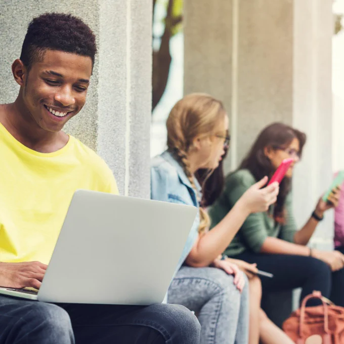 A row of diverse young people sitting outside on concrete platform using laptops and phones and smiling. 