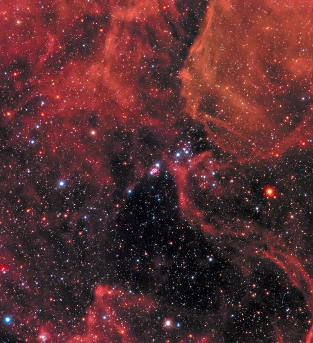 Clouds of gas and dust in shades of red cover the top of the image and a little bit of the bottom. Right in the middle is Supernova 1987a, the remnant of an exploded star. The site of the supernova is surrounded by an illuminated pinkish ring. Two faint outer rings are also visible. Stars and galaxies shine throughout, giving off red and blue light. Credit: NASA, ESA, R. Kirshner (Harvard-Smithsonian Center for Astrophysics and Gordon and Betty Moore Foundation), and M. Mutchler and R. Avila (STScI)