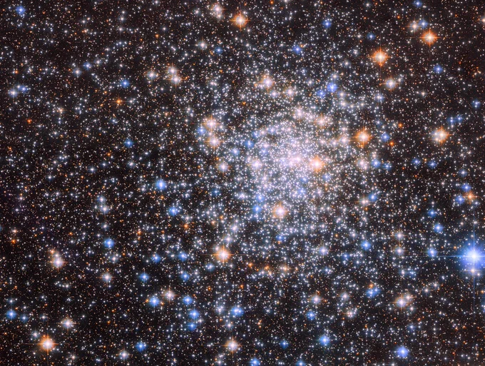 Several stars fill the image, shining against black space. The stars are more closely concentrated near the center, and more dispersed closer to the edges. These stars shine in shades of white, orange, and blue.