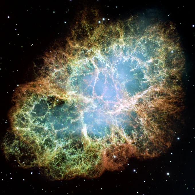 The Crab Nebula's complex oval structure lies against a black background. On the nebula’s exterior lie curtains of glowing red and orange fluffy material. Interior to this outer shell lie large-scale loops of mottled filaments of yellow-white and green. Many points of blue-white light are visible within and around the nebula.