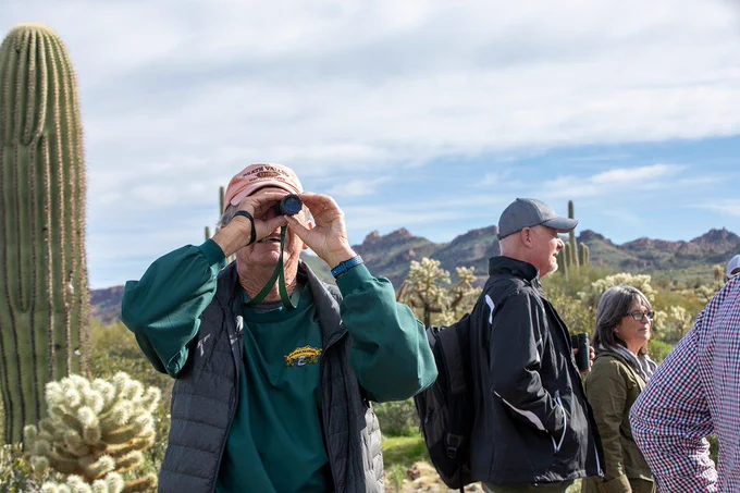 A person using a monocular to view a bird in a desert with cacti and mountains in the background, surrounded by other visitors on a bird walk.