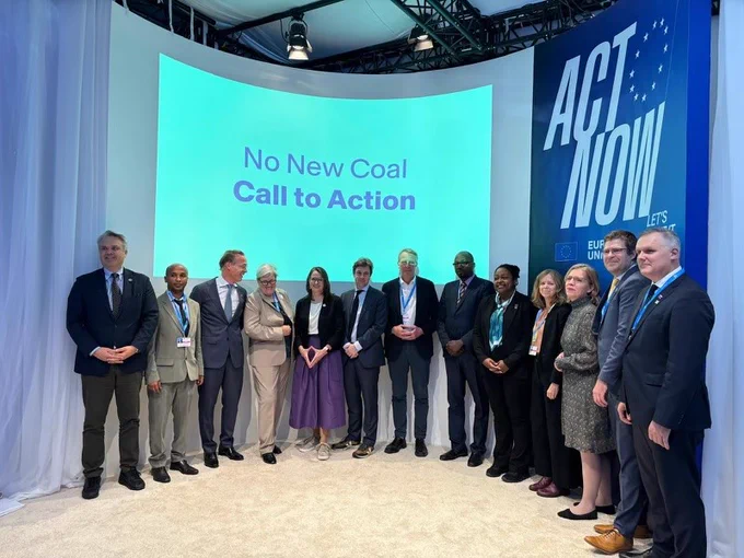 A group of individuals posing together in front of a backdrop with the words "No New Coal: Call to Action" displayed prominently. A large banner on the right side reads "ACT NOW" alongside the European Union emblem.