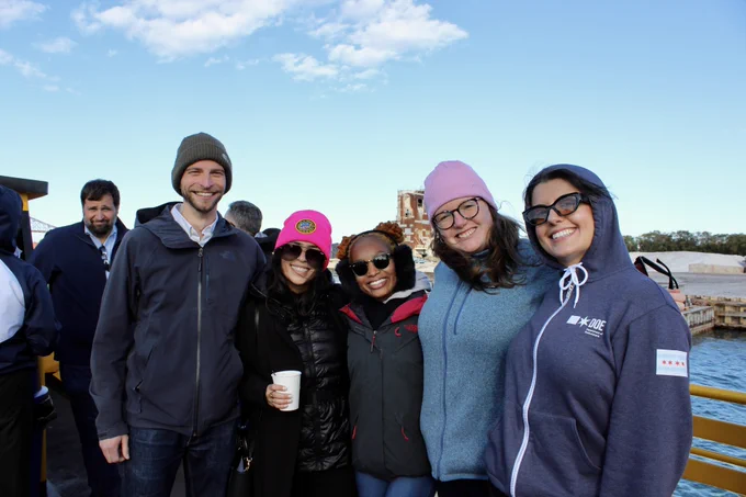 Group of five people smiling on a boat tour, with a bridge visible in the background. They are dressed in casual, warm clothing and some are wearing beanies.