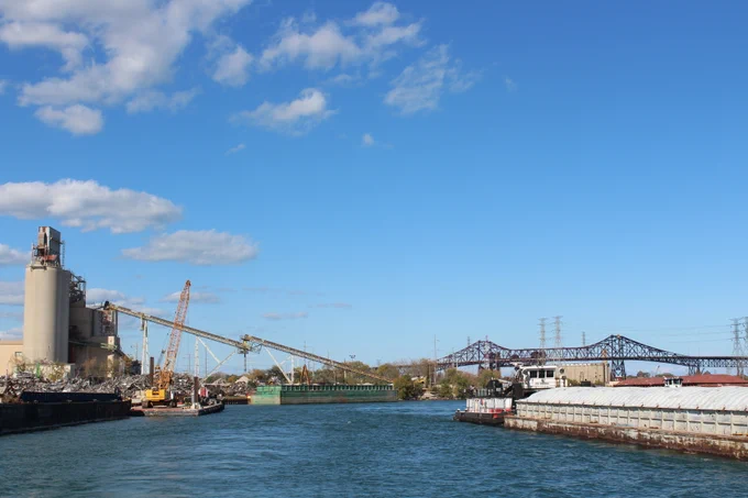 View of a serene river with industrial buildings on the left, including a large silo and a conveyor belt. Barge filled with cargo is moored in the foreground, and a steel truss bridge is visible in the distance under a clear blue sky.