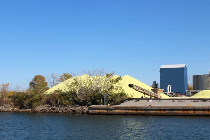 View of large yellow sulfur piles at an industrial site on the side of a river, with conveyor belts and a storage building under a clear blue sky. 