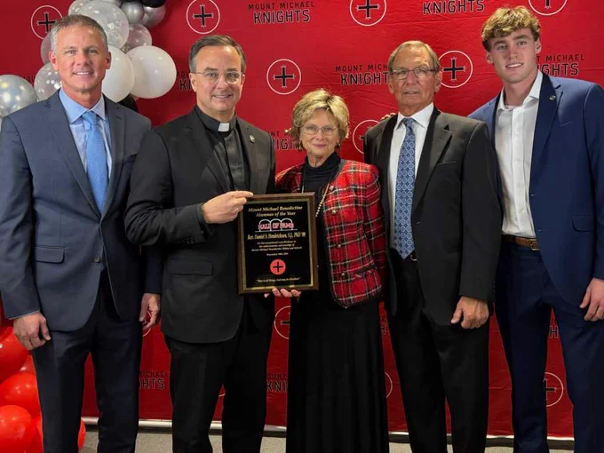 The Rev. Daniel S. Hendrickson, SJ, PhD, stands alongside his brother, mother, father, and cousin on a red backdrop at Mount Michael Benedictine High School