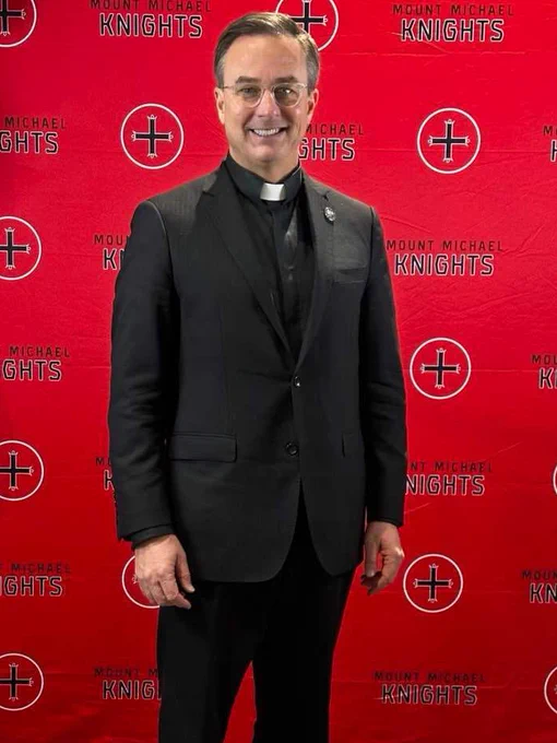 The Rev. Daniel S. Hendrickson, SJ, PhD, stands on a red backdrop at Mount Michael Benedictine High School