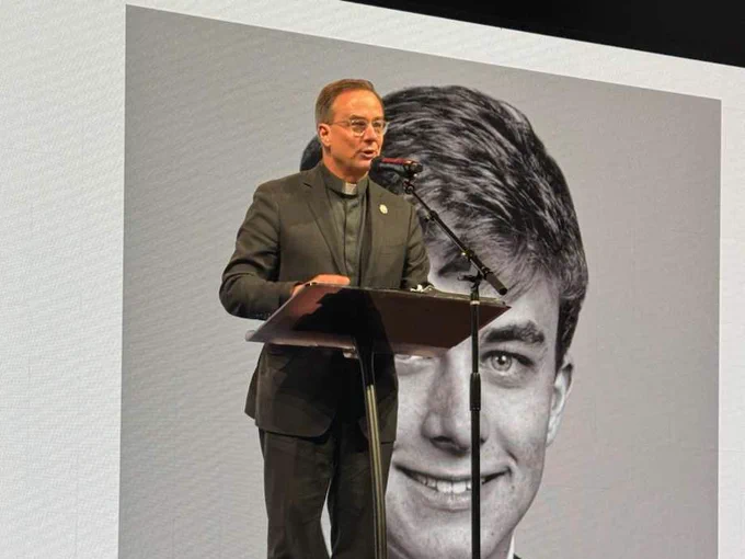 The Rev. Daniel S. Hendrickson, SJ, PhD, stands at a microphone to give a speech. A black and white photo of him as a high school student is behind him.
